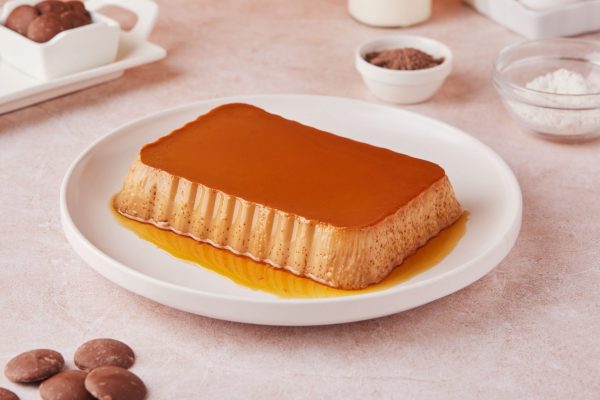 Milk Chocolate Leche Flan on a white plate, surrounded by chocolate powder, chocolate chunks, and sugar